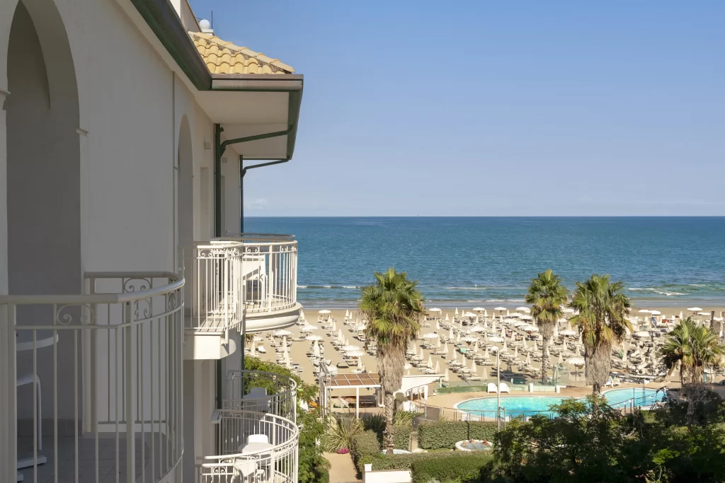 Vista dal balcone di un hotel sul mare con piscina e lettini sulla spiaggia.