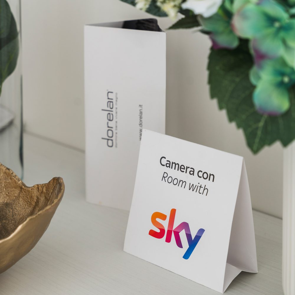 Cartello su un tavolo che pubblicizza una camera con Sky e un prosotto Dorelan, accanto a una composizione floreale.