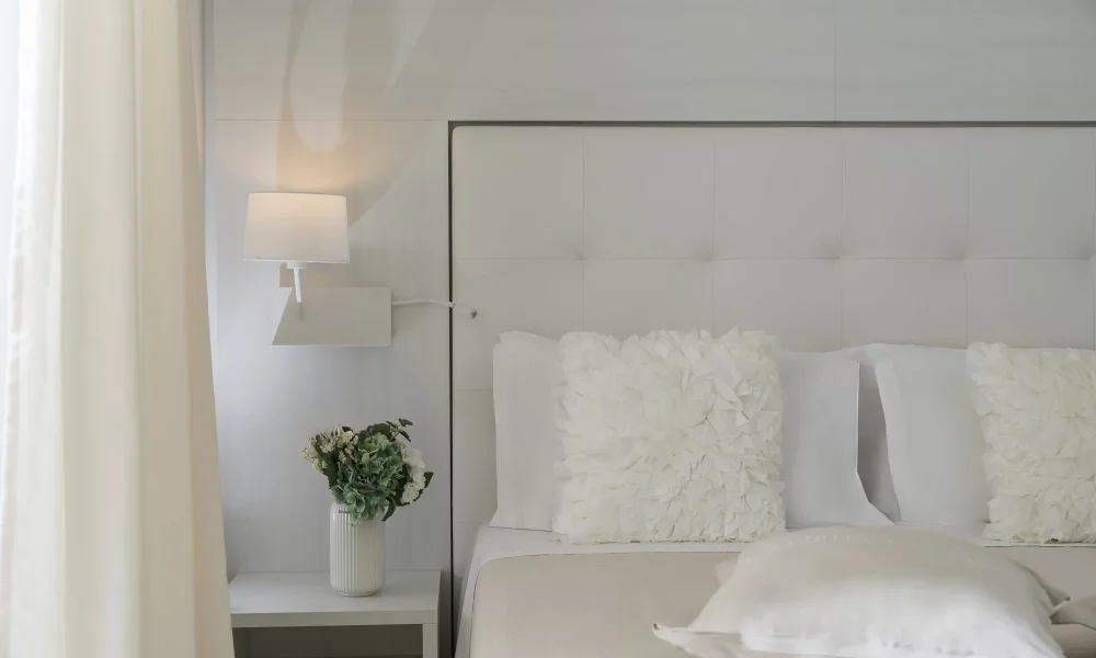 Camera da letto elegante con testiera imbottita, lampada da parete e fiore in vaso sul comodino.
