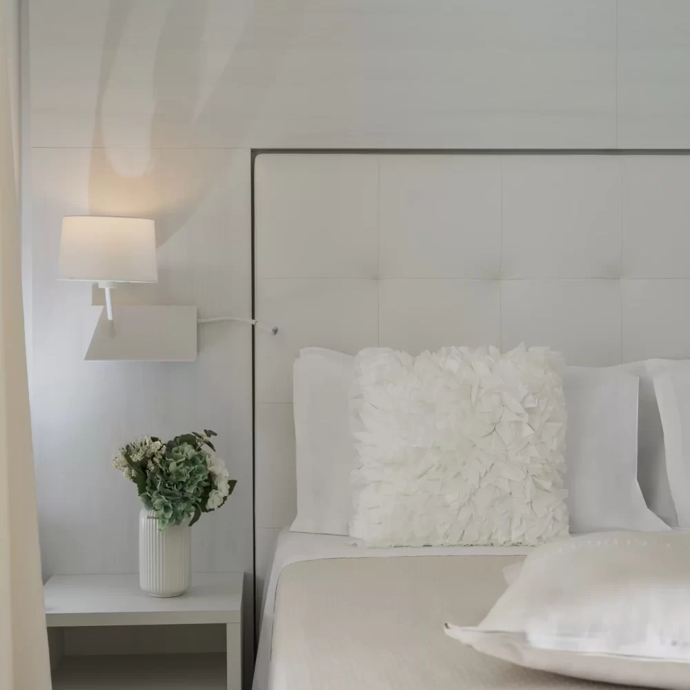 Camera da letto elegante con testiera imbottita, lampada da parete e fiore in vaso sul comodino.