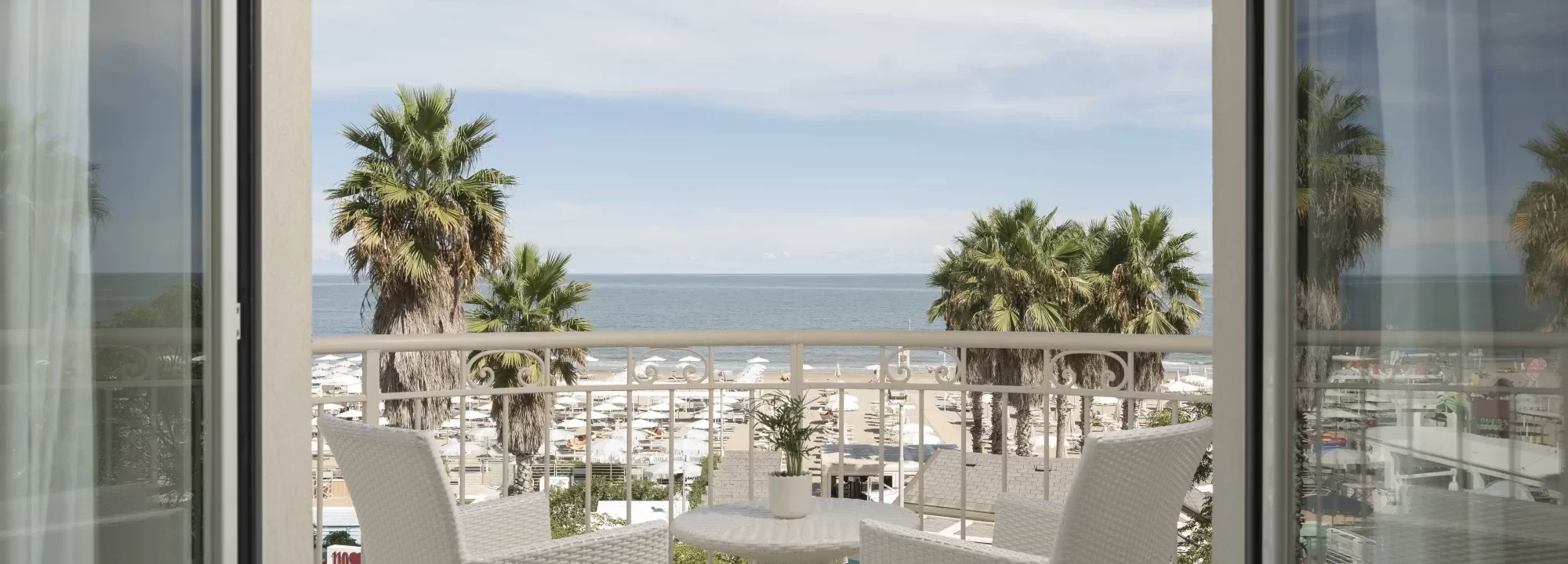 Blick von einem Balkon auf das Meer und den Strand mit Palmen und weißen Möbeln im Vordergrund.