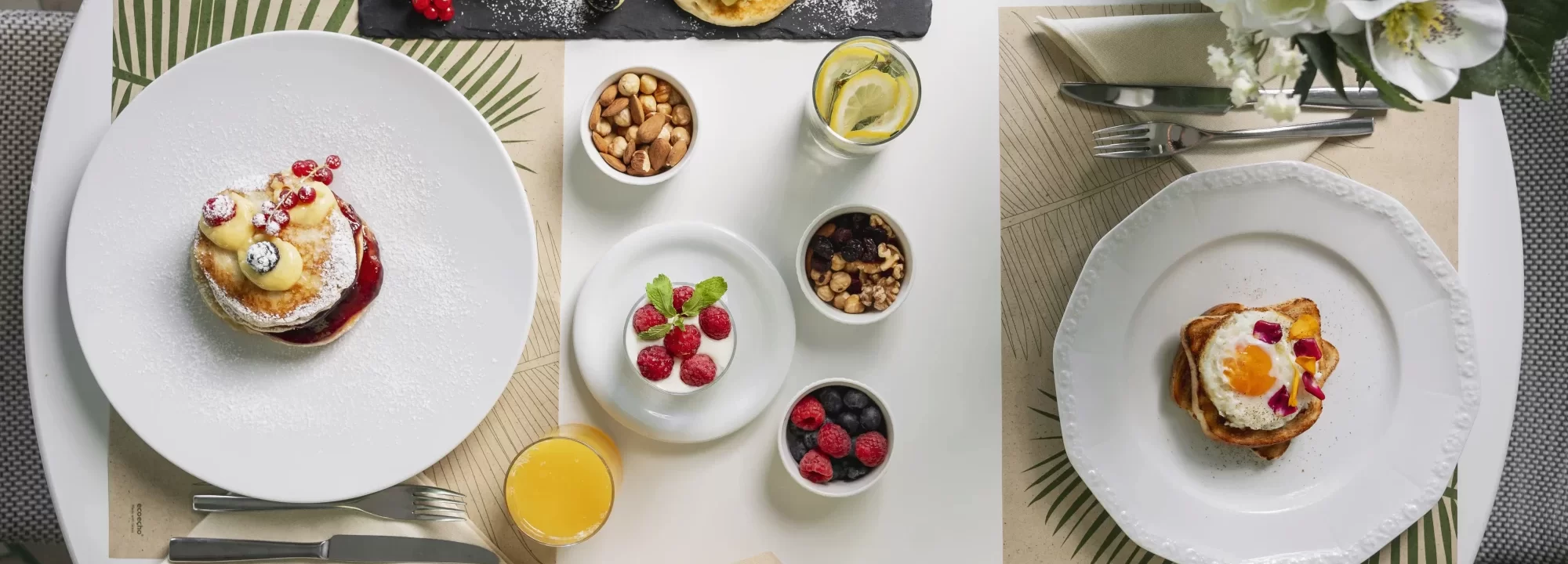 Colazione gourmet con pancake, croissant, toast, frutta fresca e bevande su un tavolo elegantemente apparecchiato.