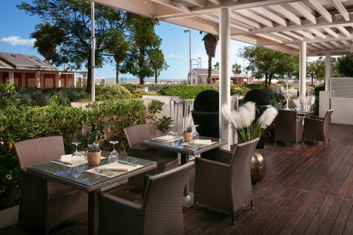 Terrazza ristorante con tavoli apparecchiati e vista sul mare circondata da vegetazione.