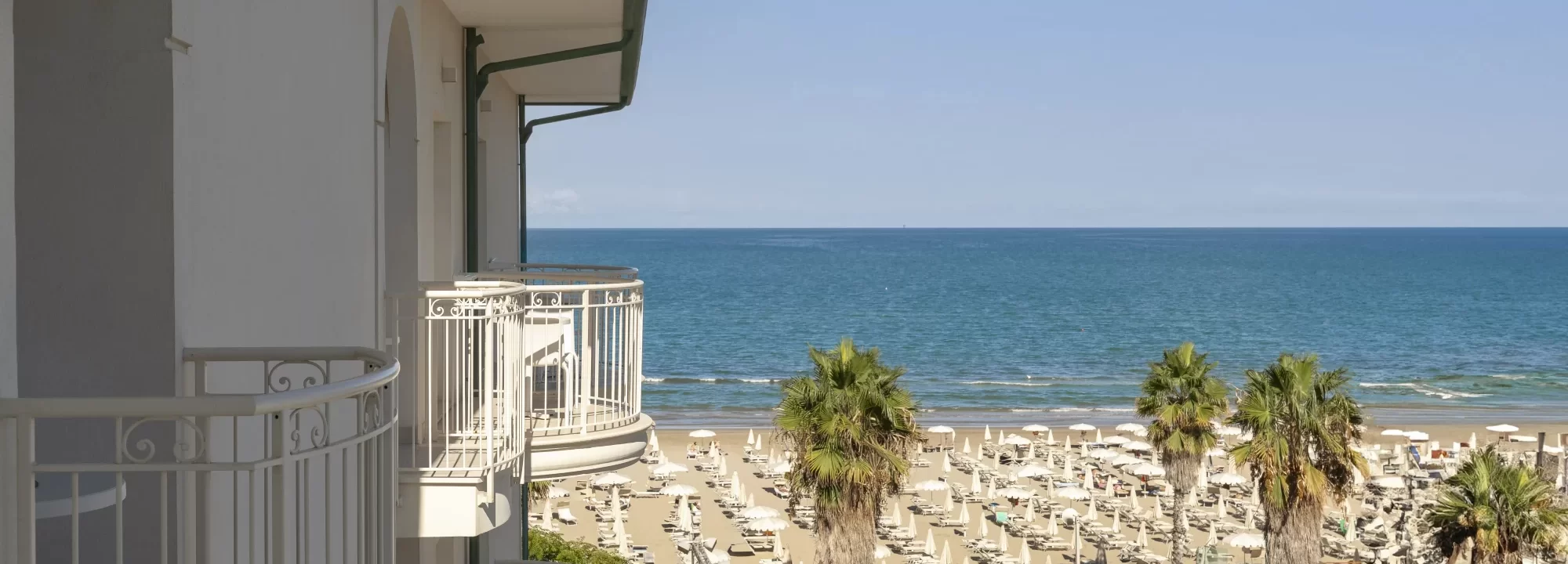 Vista dal balcone di un hotel sul mare con piscina e lettini sulla spiaggia.