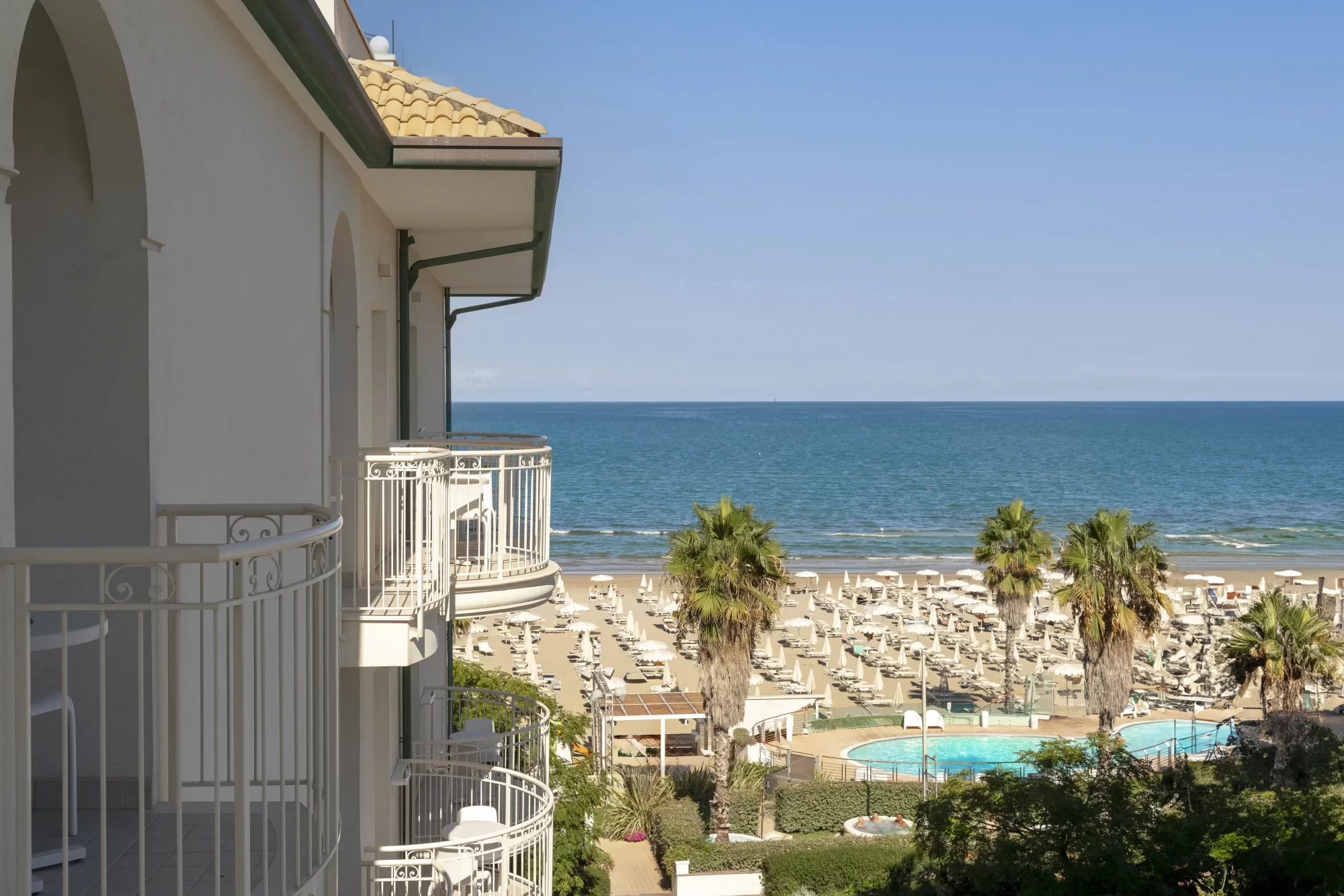 Vista dal balcone di un hotel sul mare con piscina e lettini sulla spiaggia.