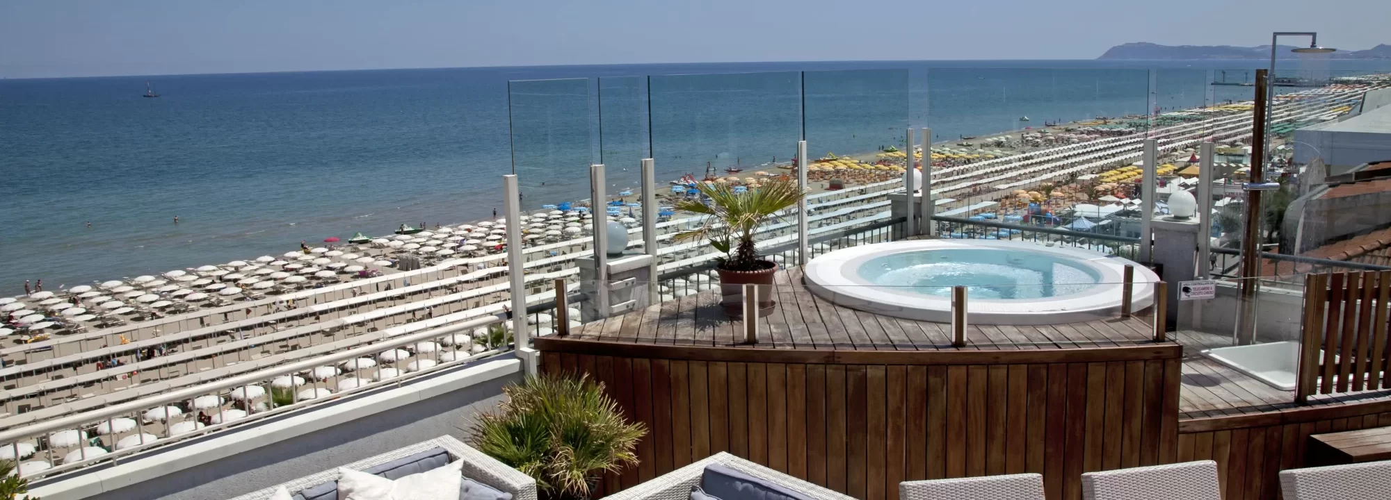 Dachterrasse mit Whirlpool und Blick auf einen Strand mit vielen Sonnenschirmen.