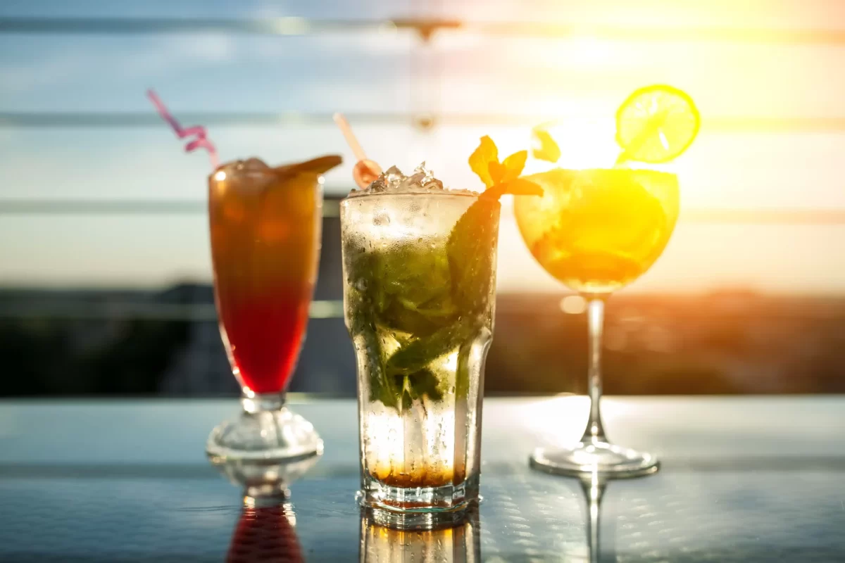 Tre cocktail colorati su un tavolo al tramonto con riflessi di luce.