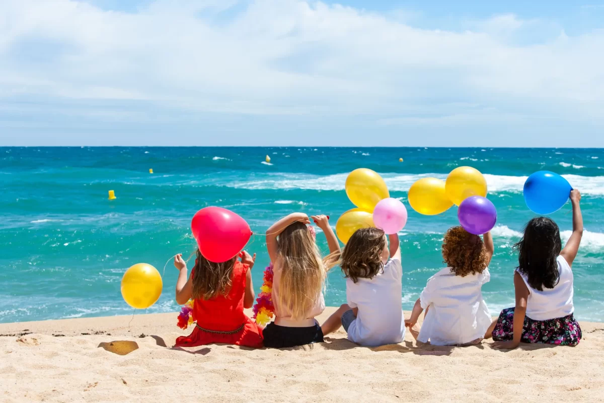 Bambini seduti sulla spiaggia che tengono palloncini colorati di fronte al mare.