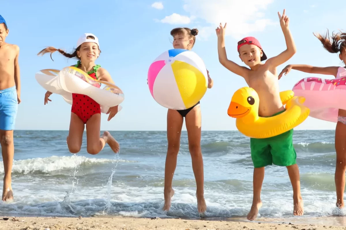 Bambini che saltano e giocano sulla spiaggia con salvagenti e palloni colorati.