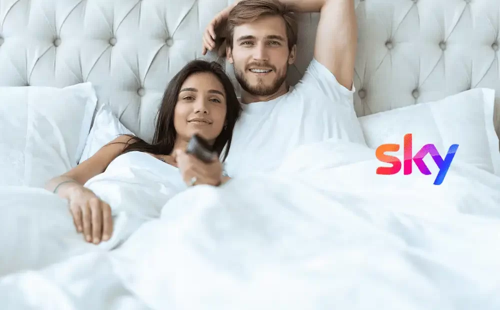 Coppia felice a letto guarda la televisione con il logo Sky visibile.