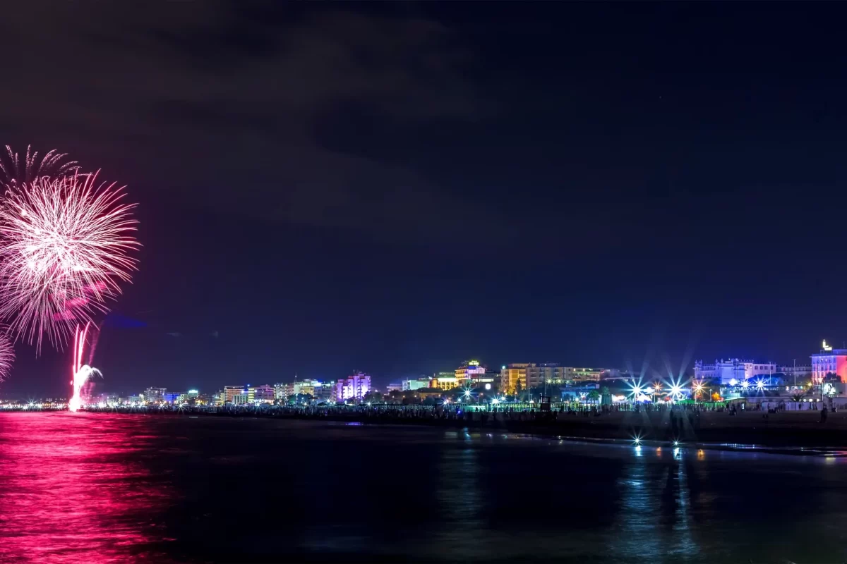 Spettacolo pirotecnico sul mare di notte con una città illuminata sullo sfondo.