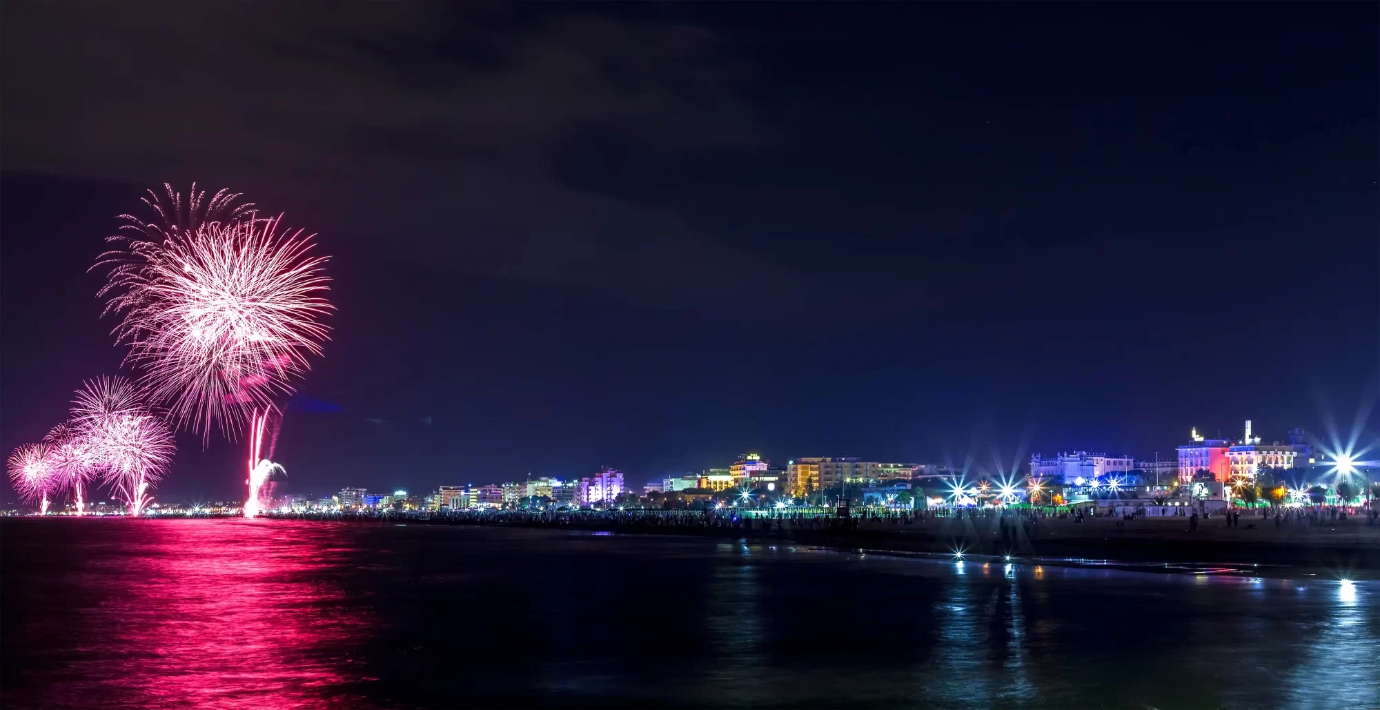 Spettacolo pirotecnico sul mare di notte con una città illuminata sullo sfondo.