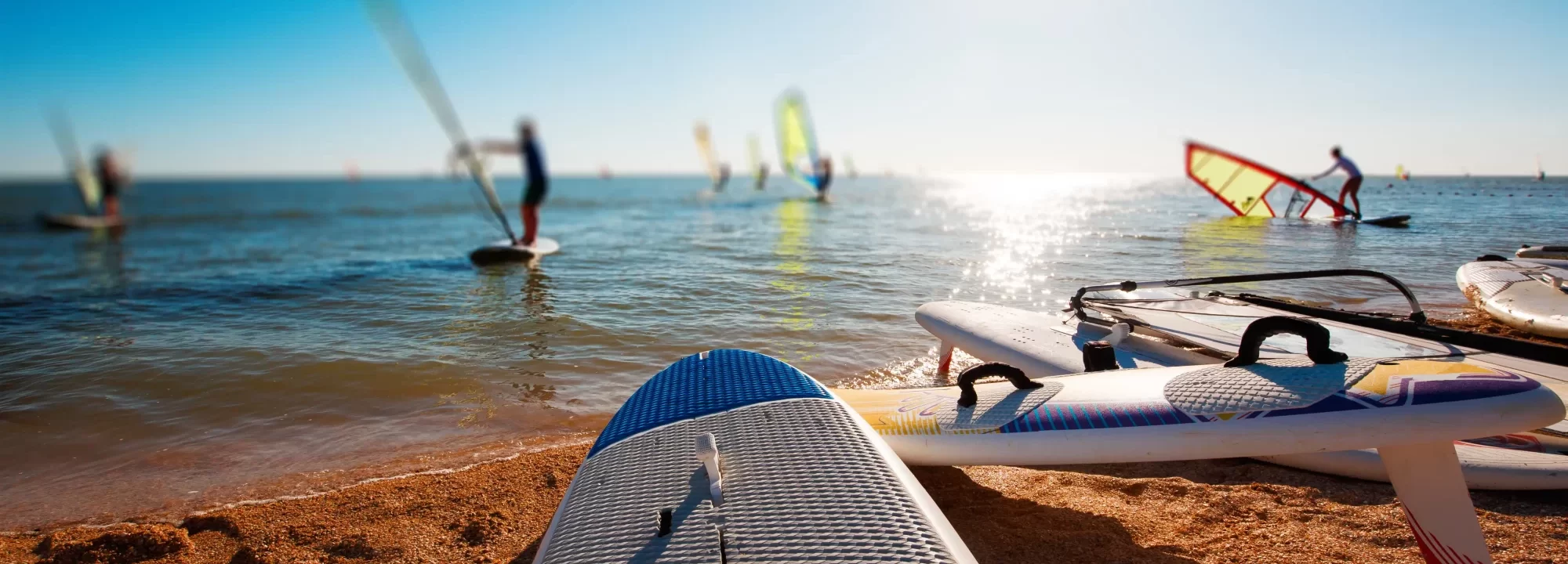 Tavole da windsurf sulla spiaggia con persone che praticano windsurf al largo.