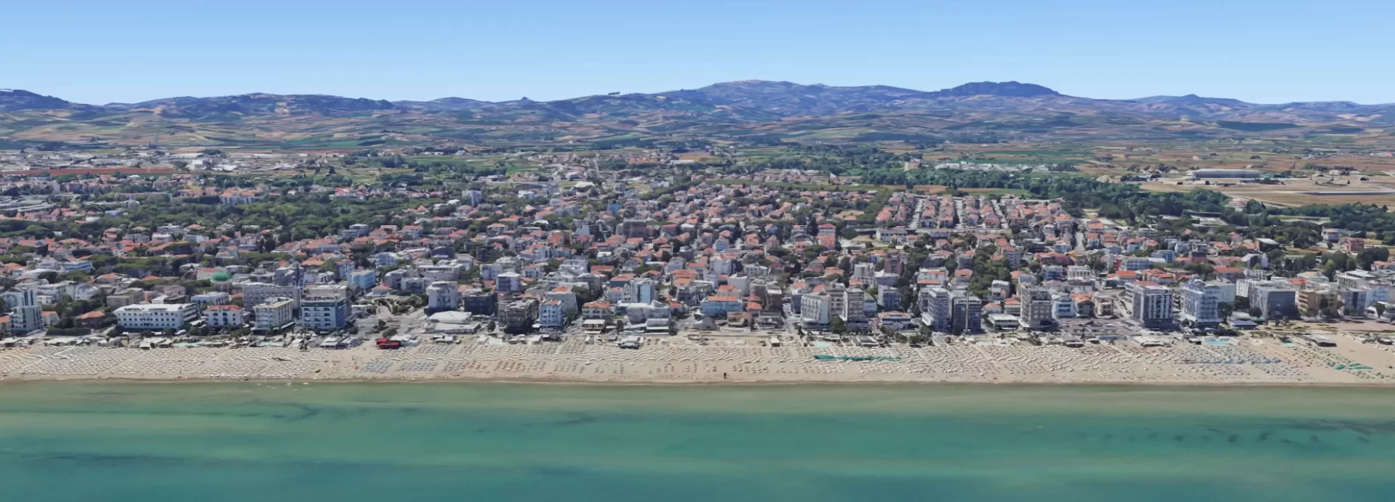 Veduta panoramica di una città costiera con spiaggia affollata e colline sullo sfondo.