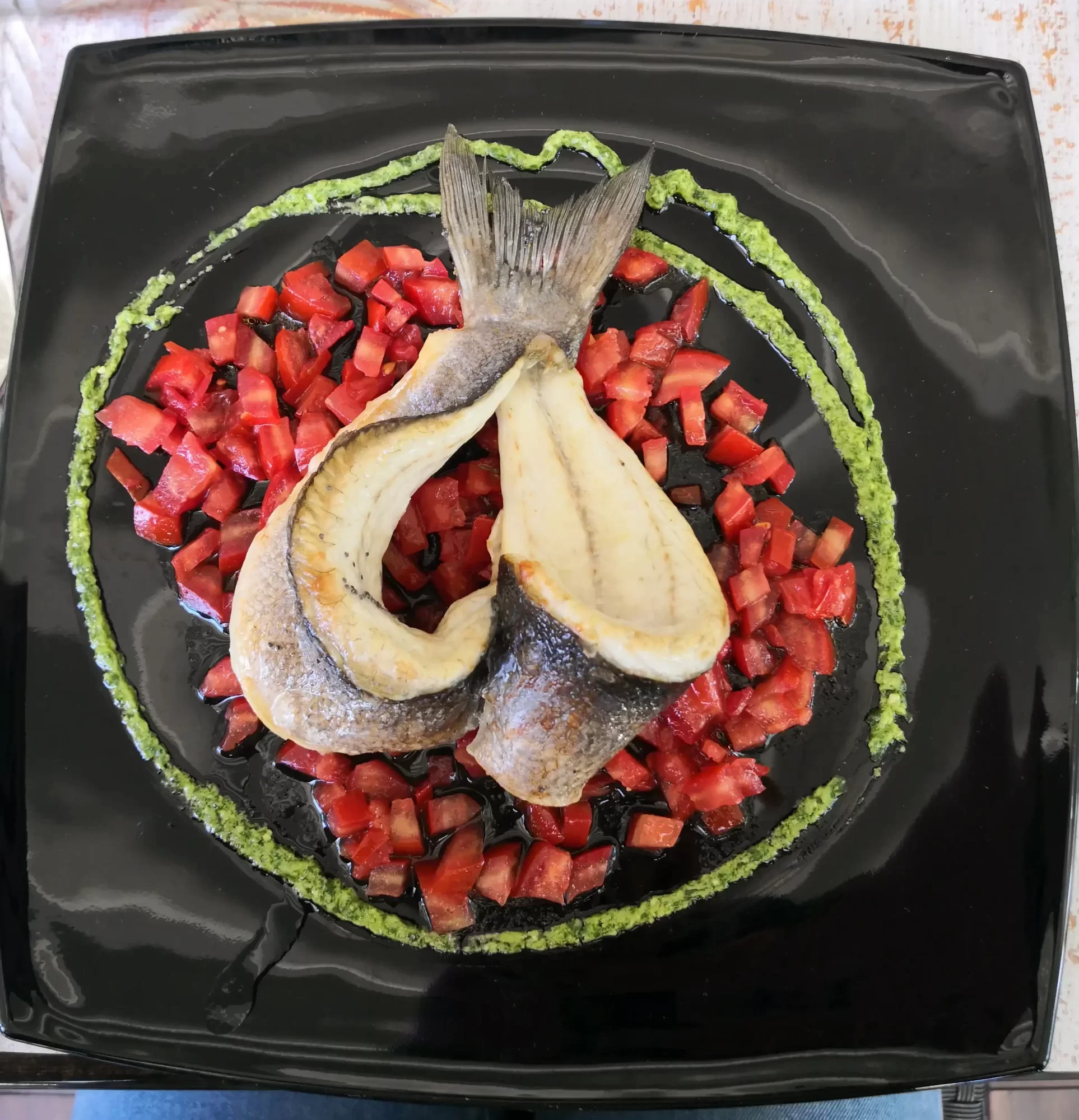 Un elegante piatto di pesce con il corpo disposto a cerchio su una salsa verde e guarnito con cubetti di pomodoro.